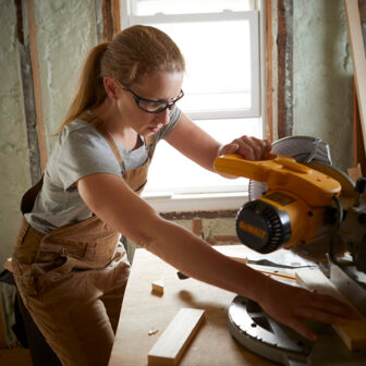 Female contractor using a miter saw, wearing safety googles and coveralls.