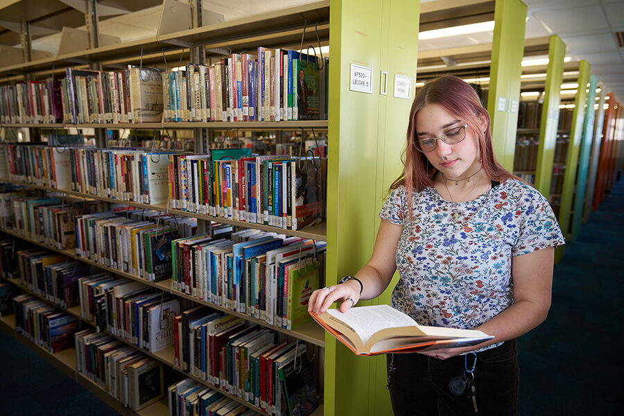 Female student standing in library stacks looking at a book