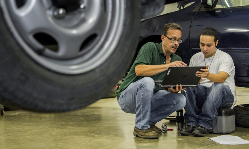 Caucasian male instructor and Latin American, male student knelt down reviewing a laptop in the automotive technology lab