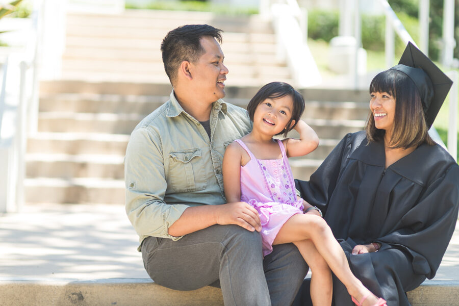 Father, young daughter, and mother sitting outside. Mother is wearing a black, graduation cap and gown