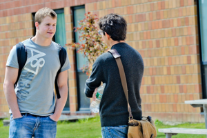 Two young, white males interact outside on campus in front of a brick building.