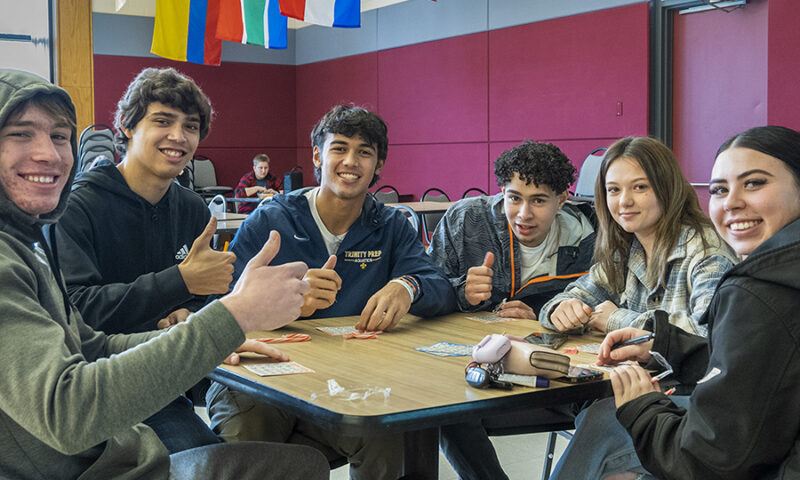 Group of students sitting around table in the cafeteria playing bingo, giving a thumbs up