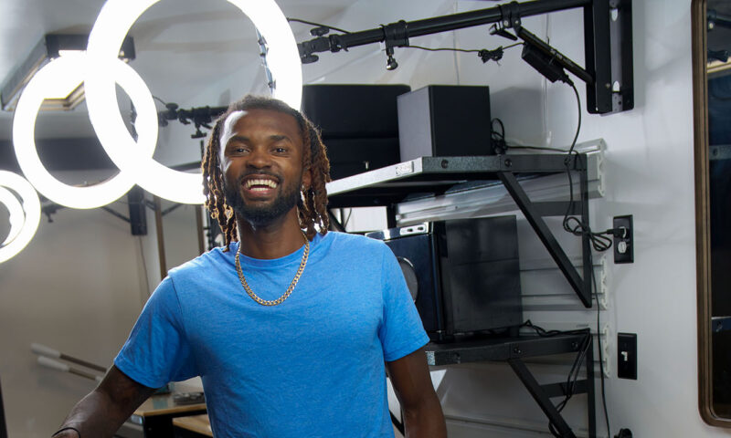 African American male with dreadlocks smiling wearing a royal blue t-shirt and gold chain.