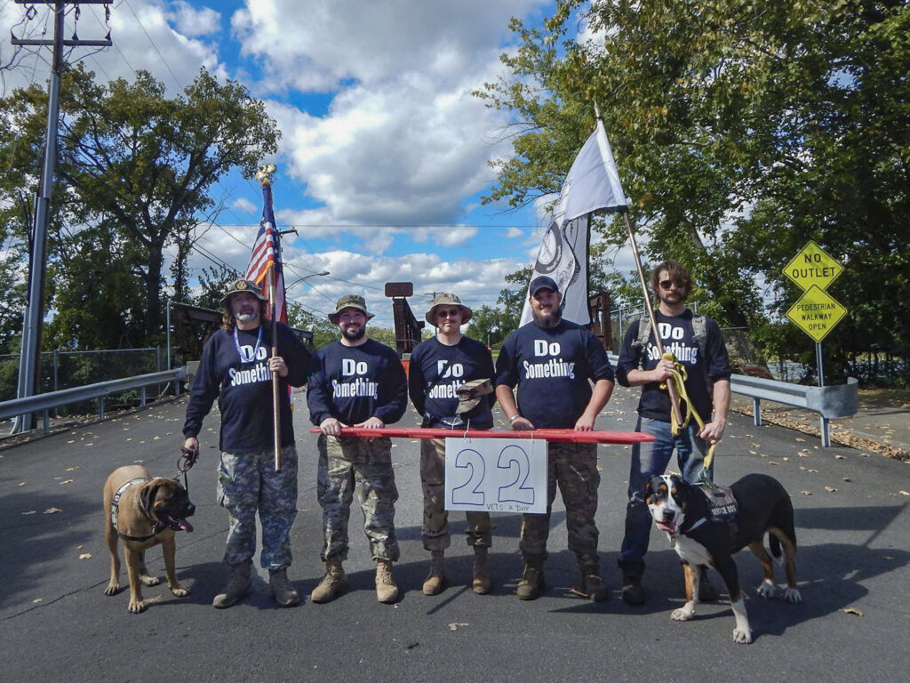 Five caucasian men wearing blue shits that read "Do Something". They are holding a banner that says 22 push up challenge. Two service dogs bookend the men